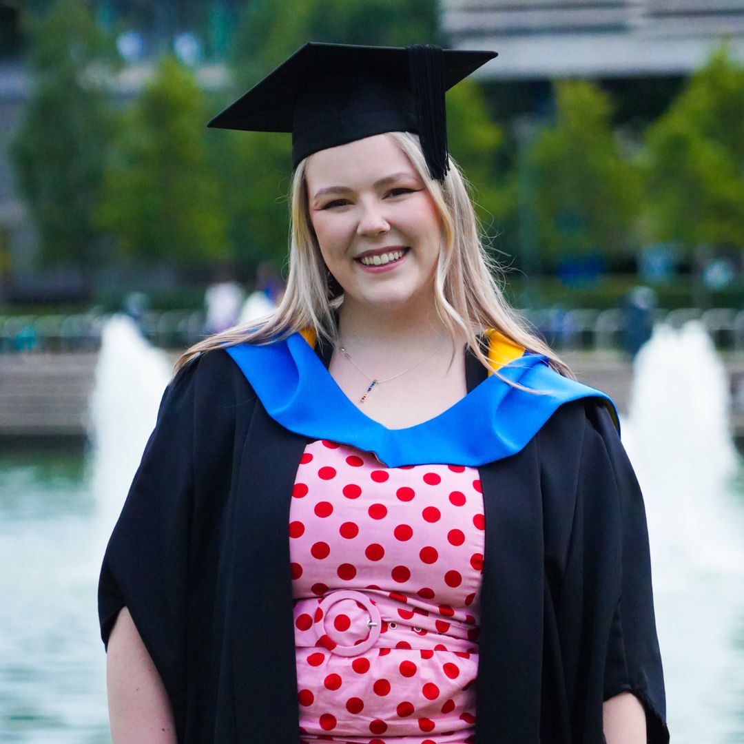 Niamh Scully wears a graduation gown and cap in front of the UCD lake.