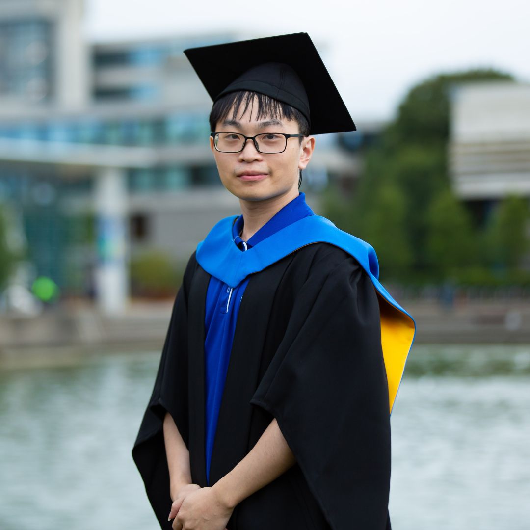 Chen Chen in graduation robes by the main lake at UCD.